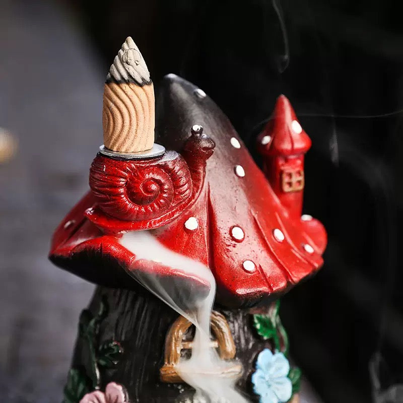 Front view of the whimsical red mushroom fairy house backflow incense burner on a bright white background. Displays the soothing smoke waterfall effect cascading from the snail on the red spotted roof down over the intricate stone doorway and colorful floral details. Perfect for relaxing home decor.