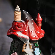 Front view of the whimsical red mushroom fairy house backflow incense burner on a bright white background. Displays the soothing smoke waterfall effect cascading from the snail on the red spotted roof down over the intricate stone doorway and colorful floral details. Perfect for relaxing home decor.
