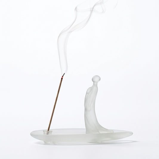 Frosted glass incense holder with a minimalist human figure standing on a translucent oval base, holding a lit incense stick with soft rising smoke on a clean white background.