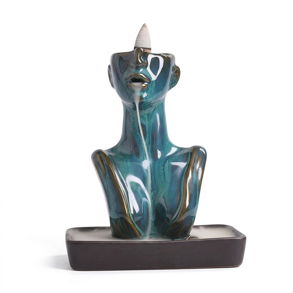 Glossy glazed blue abstract face backflow incense burner with a cone incense placed on top, white smoke flowing from the lips into a rectangular base tray on a light background.