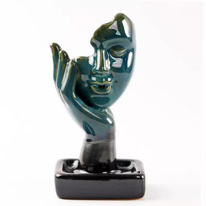 A glossy blue-green ceramic abstract face incense burner with a sculpted hand detail, displayed on a black rectangular base with built-in holders for a backflow cone and incense sticks.