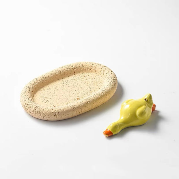 A studio shot of a glossy yellow ceramic duck figurine lying next to an oval, beige tray with a rough, porous stone-like texture. The set is displayed on a plain white background.