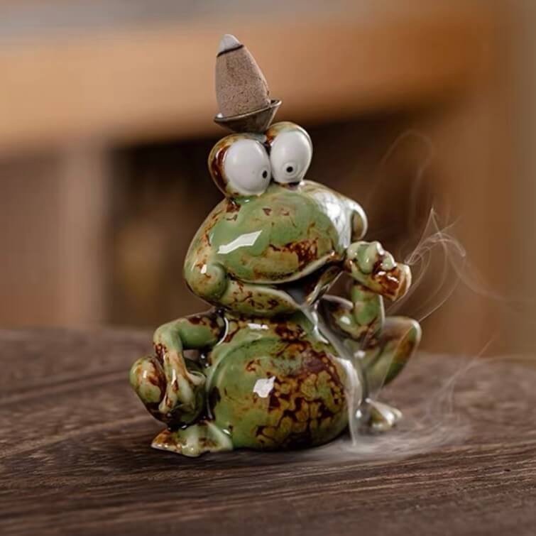 A close-up of a glossy green speckled ceramic backflow incense burner shaped like a frog sitting on a wooden surface, featuring a lit incense cone on its head with smoke gently flowing downwards from its mouth.