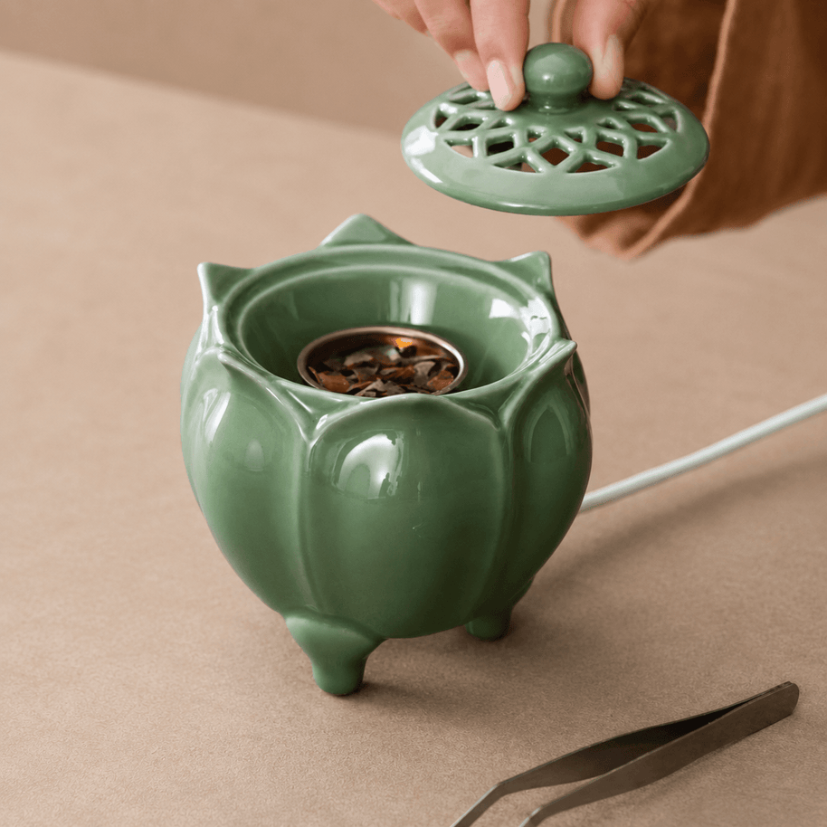 Hand lifting the vented lid from a green lotus shaped electric incense burner showing the metal incense dish inside