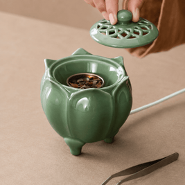 Hand lifting the vented lid from a green lotus shaped electric incense burner showing the metal incense dish inside