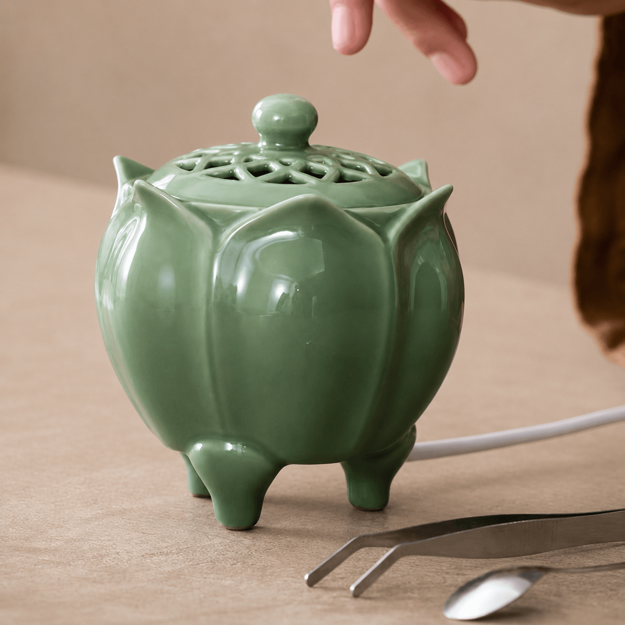 Green lotus shaped electric incense burner with a decorative vented lid displayed beside a metal spoon and tweezers on a beige surface