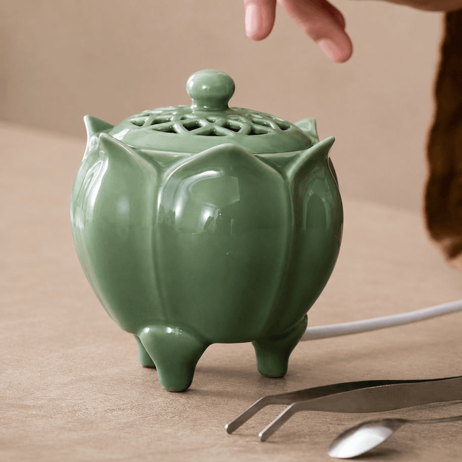 Green lotus shaped electric incense burner with a decorative vented lid displayed beside a metal spoon and tweezers on a beige surface