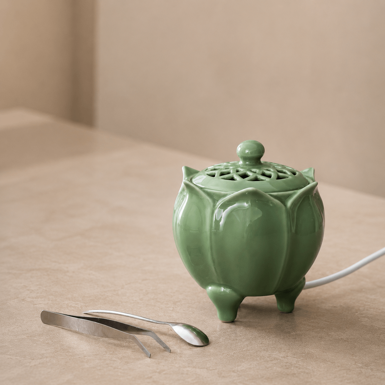 Green lotus shaped electric incense burner displayed on a beige surface with a metal spoon and tweezers beside it