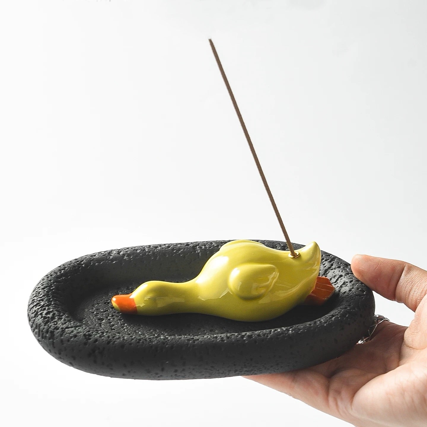 A hand holding the assembled incense set, featuring a glossy yellow ceramic duck resting on a black, porous oval tray. An incense stick is inserted into the duck, illustrating the product's size and usage.