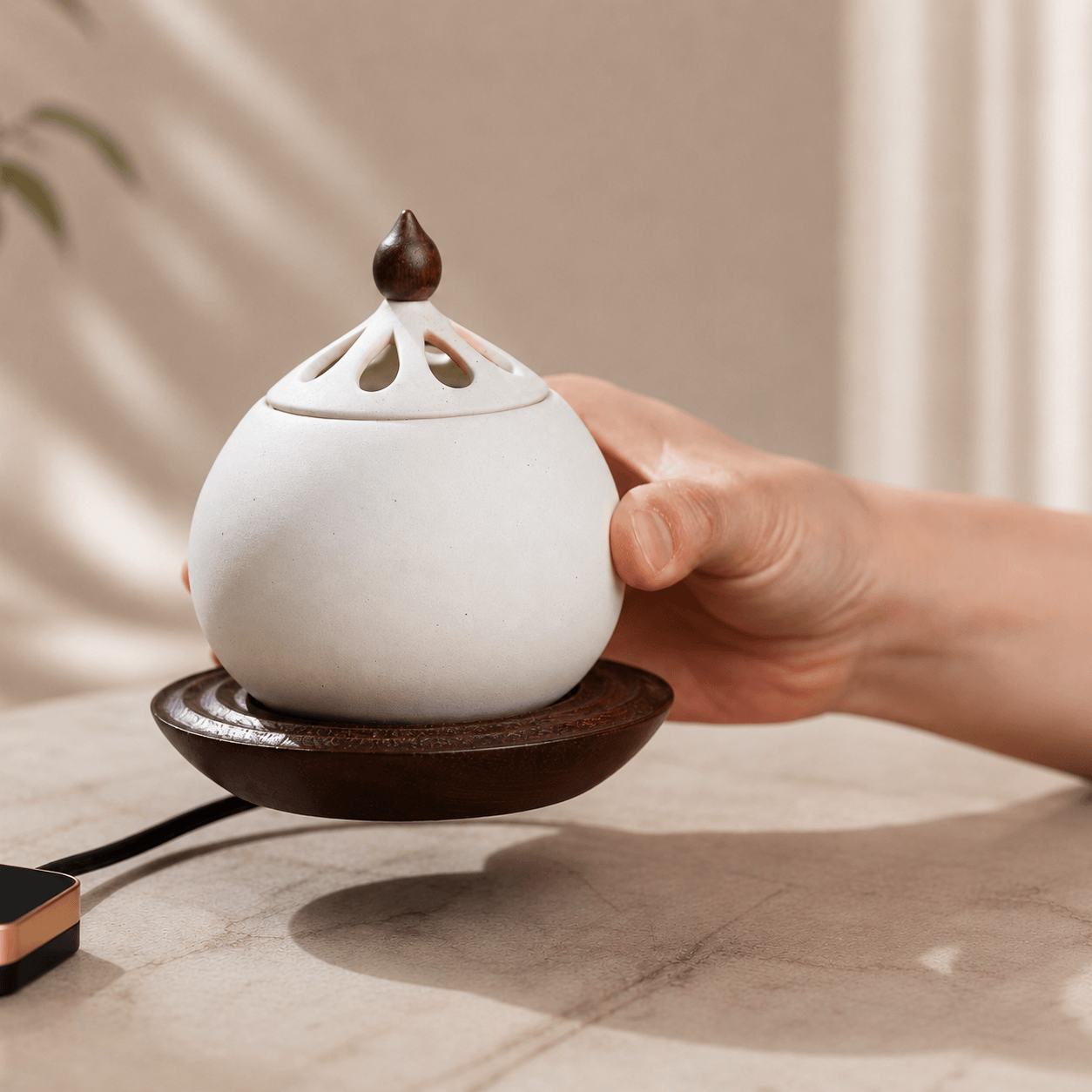 A hand gently touching the side of a white electric ceramic incense burner resting on a solid wood base, placed on a light-colored table.