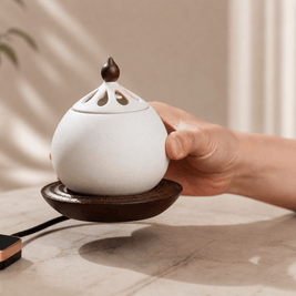 A hand gently touching the side of a white electric ceramic incense burner resting on a solid wood base, placed on a light-colored table.