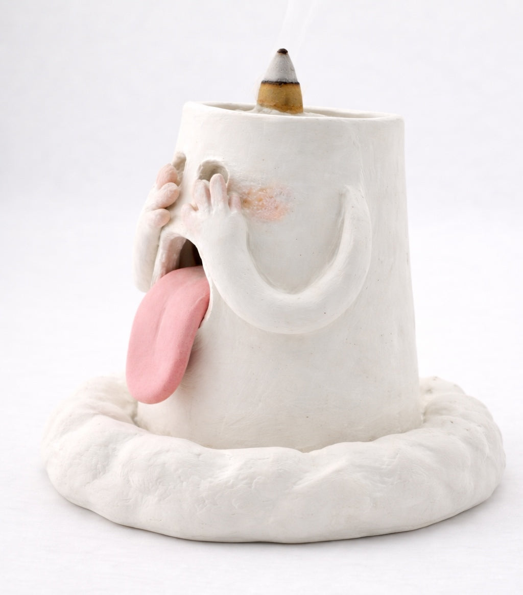 Side view of a handcrafted blushing ghost backflow incense burner with hands over its eyes, pink tongue out, and an incense cone on top against a white background