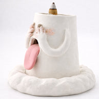 Side view of a handcrafted blushing ghost backflow incense burner with hands over its eyes, pink tongue out, and an incense cone on top against a white background