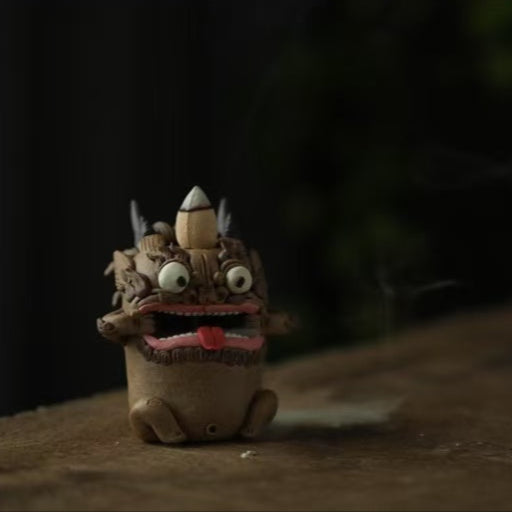 Angled side view of a handcrafted purple clay guardian backflow incense burner with prominent horns and wide-open mouth, red tongue exposed and white mist flowing from its mouth onto a wooden surface.