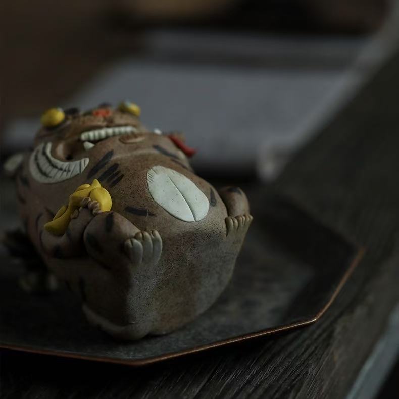 Handcrafted purple clay tiger guardian incense burner shown reclining on a dark tray, highlighting its rounded belly, tiny paws, yellow ingot detail, striped body, and playful handmade ceramic character in a cozy tabletop setting.