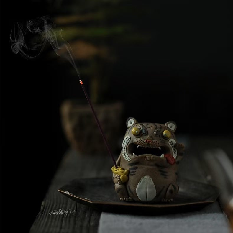 Handcrafted purple clay tiger guardian incense burner displayed on a dark tray with a lit stick incense, featuring glowing smoke, bulging yellow eyes, a red tongue, and gold ingot detail in a moody tabletop setting.