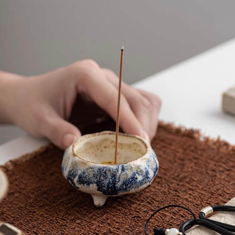 Close up of a handmade blue marbled ceramic incense holder with an upright incense stick while a hand steadies the bowl