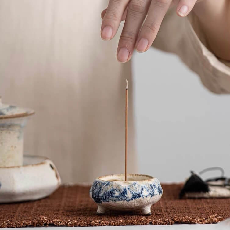 Hand hovering above a burning incense stick placed in a handmade blue marbled ceramic incense holder on a woven mat