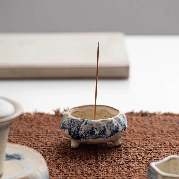 Handmade blue marbled ceramic incense holder with three short feet holding a single upright incense stick on a woven mat
