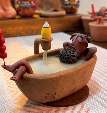 Handmade ceramic backflow incense burner featuring The Relaxing Sage relaxing in a bathtub with an incense cone and flowing smoke in a warm cozy interior setting