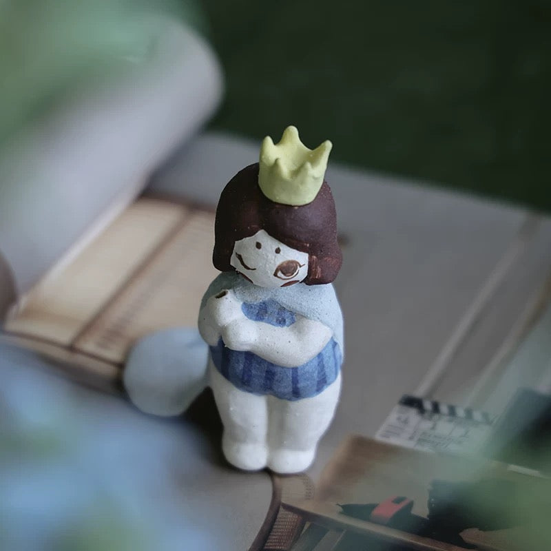 Single handmade ceramic girl incense holder with a yellow crown and blue outfit, standing on books with a leaf-shaped tray behind.