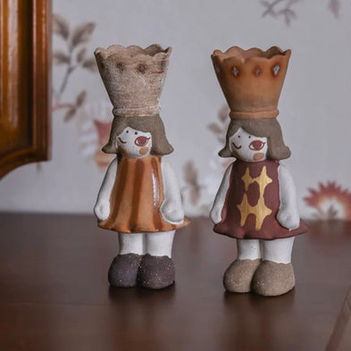 Two handmade ceramic girl incense burners with crown tops and patterned dresses standing on a wooden surface with a floral backdrop.