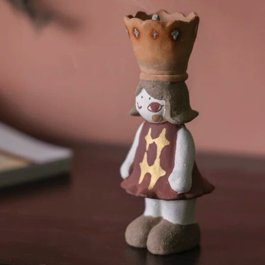 Side view of a handmade ceramic girl figurine incense burner with a crown-shaped cone holder on top and a star-pattern dress, on a wooden surface with a warm backdrop.