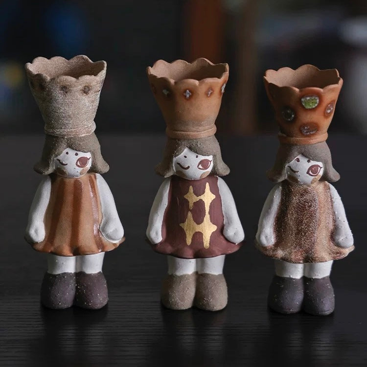 Three handmade ceramic girl incense burners in a row with crown-shaped cone holders and different dress patterns on a dark tabletop.