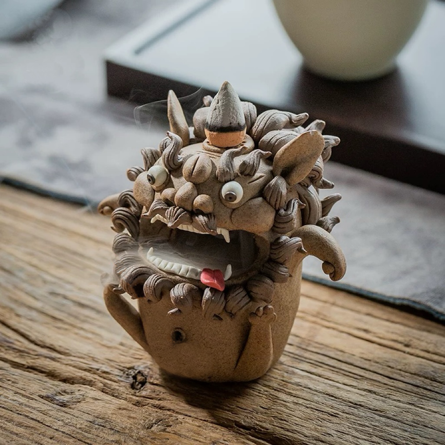Angled view of a handmade ceramic guardian beast backflow incense burner with an incense cone on top, bulging eyes, fangs, curled mane, and drifting smoke on a rustic wooden surface