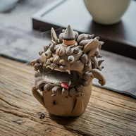Angled view of a handmade ceramic guardian beast backflow incense burner with an incense cone on top, bulging eyes, fangs, curled mane, and drifting smoke on a rustic wooden surface
