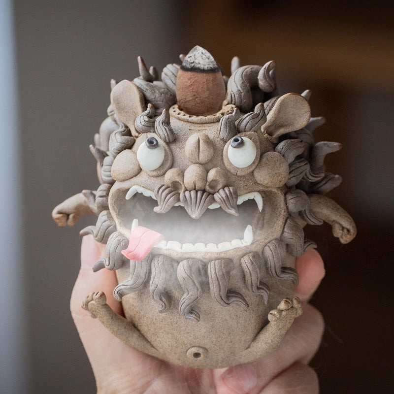 Handmade ceramic guardian beast backflow incense burner shown held in hand, with bulging eyes, curled mane, fangs, tongue out, and incense smoke flowing from the mouth