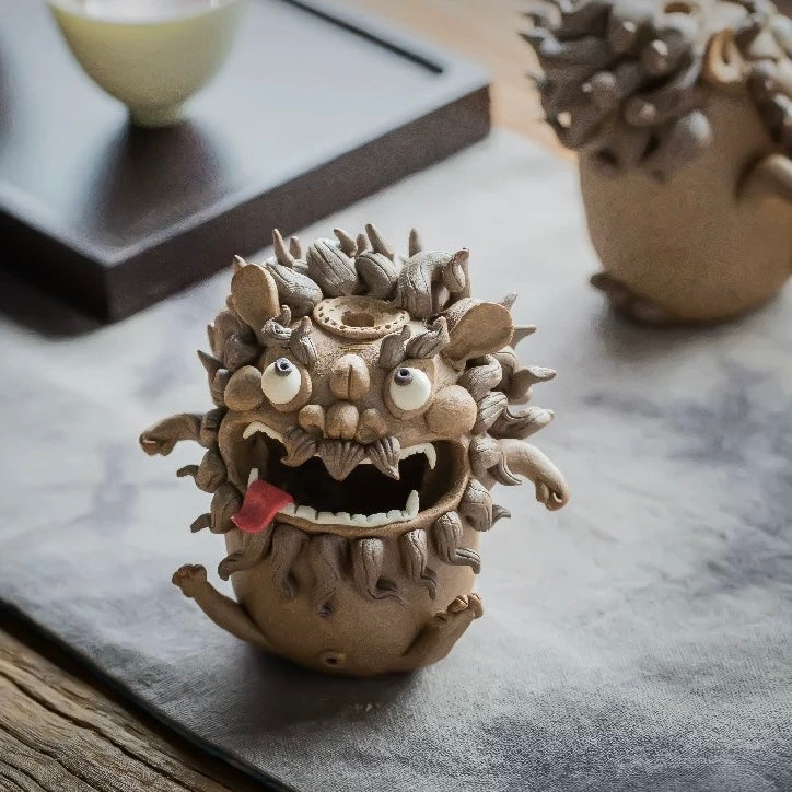 Handmade ceramic guardian beast backflow incense burner with bulging eyes, fangs, curled mane, and tongue out on a tabletop in a warm interior setting
