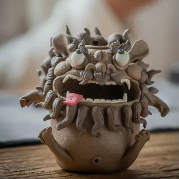 Handmade ceramic guardian beast backflow incense burner with bulging eyes, curled mane, fangs, and tongue out displayed on a wooden surface in a warm rustic setting