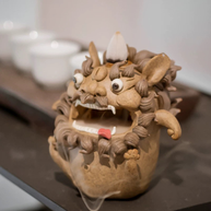 Handmade ceramic guardian beast backflow incense burner with an incense cone on top, bulging eyes, fangs, curled mane, and flowing smoke on a tabletop in a warm indoor setting