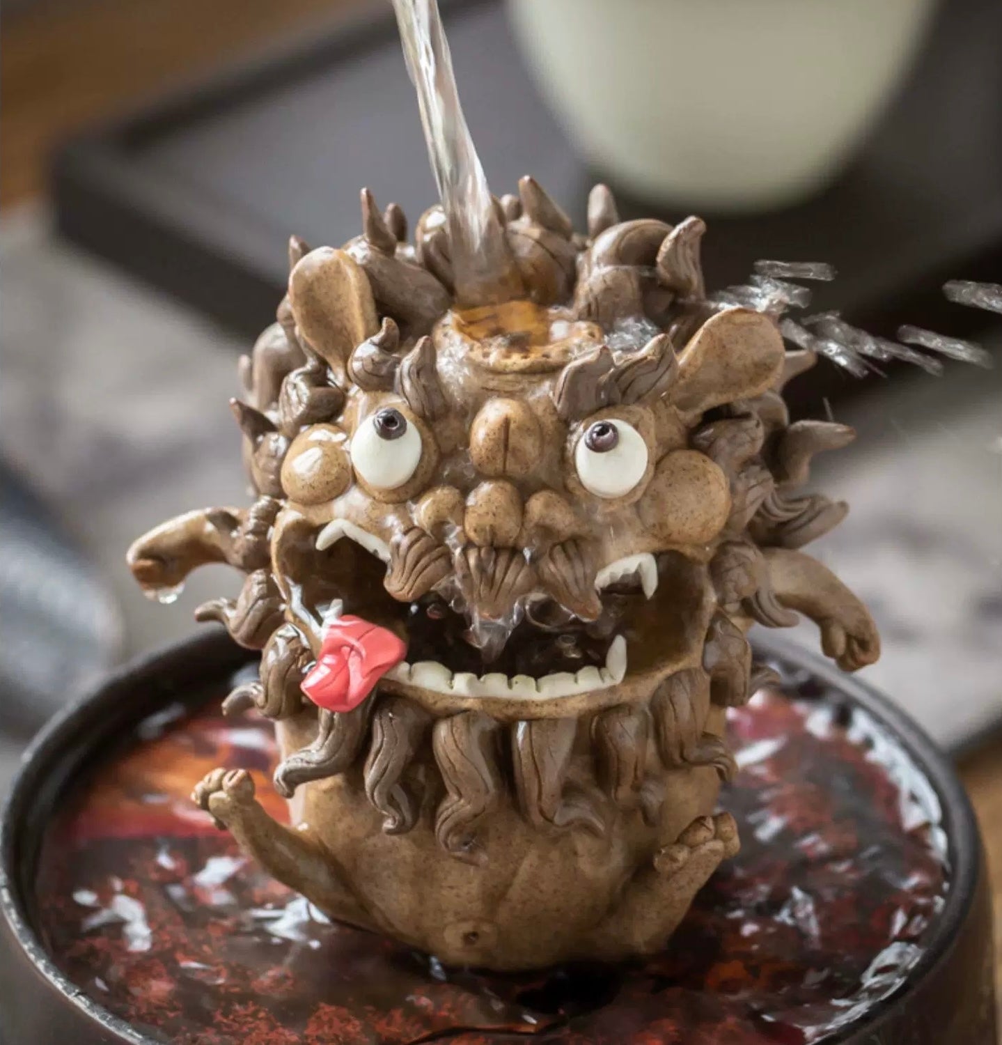  Handmade ceramic guardian beast backflow incense burner with bulging eyes, fangs, curled mane, and tongue out, shown with water flowing into the top opening on a decorative tray
