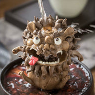  Handmade ceramic guardian beast backflow incense burner with bulging eyes, fangs, curled mane, and tongue out, shown with water flowing into the top opening on a decorative tray