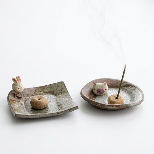 Handmade ceramic incense holder set displayed on a rustic wooden table, featuring a rabbit figurine on a square dish and a pig figurine on a round dish with a lit incense stick, beside an open book.