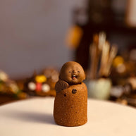 Close-up of The Joyful Little Monk handmade ceramic incense stick holder with a smiling face and fan on a white tabletop in a warm blurred interior setting