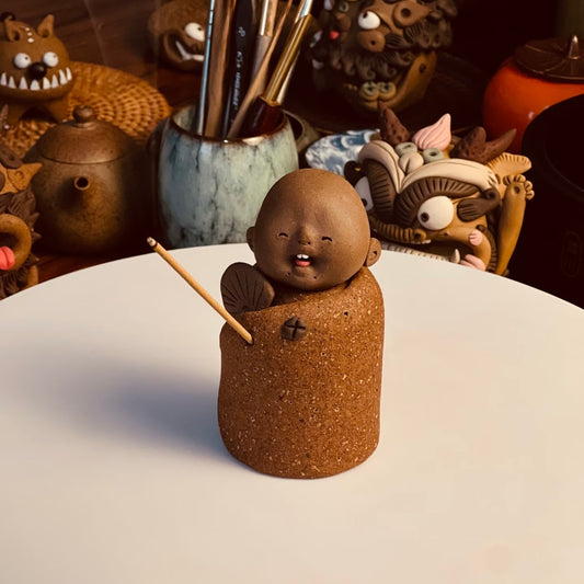 Handmade ceramic incense stick holder shaped like The Joyful Little Monk displayed with an incense stick on a white tabletop in a warm decorative setting