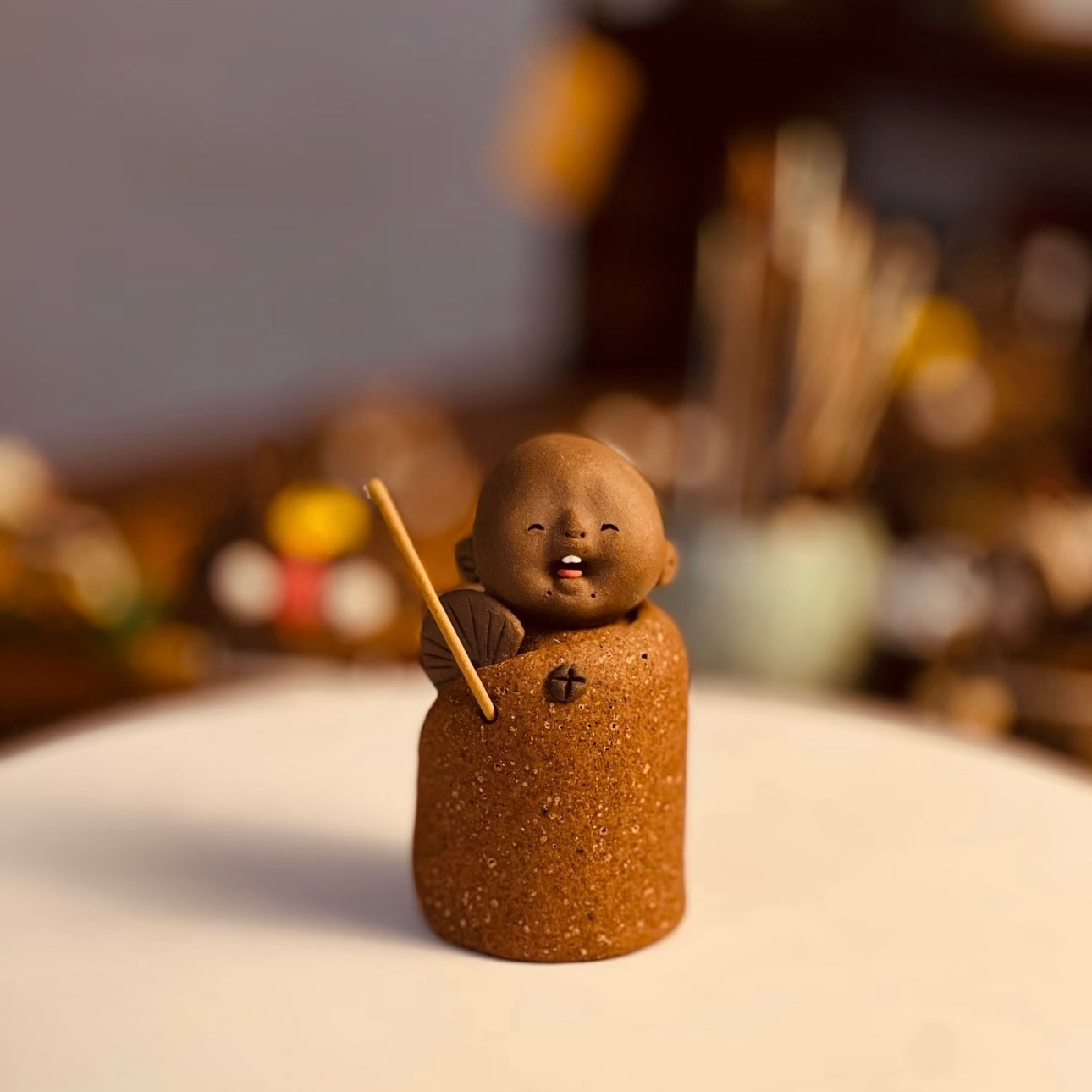 Handmade ceramic incense stick holder shaped like The Joyful Little Monk holding a fan and an incense stick on a white surface