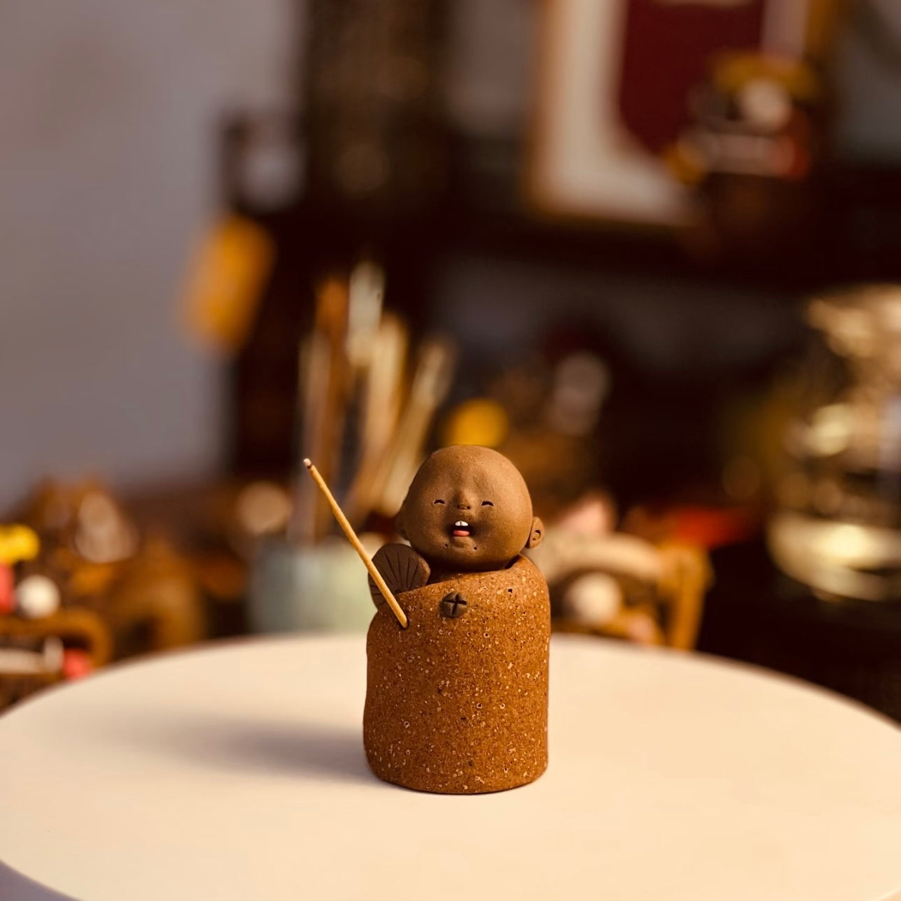 Handmade ceramic incense stick holder shaped like The Joyful Little Monk with an incense stick on a white tabletop in a warm softly blurred interior setting
