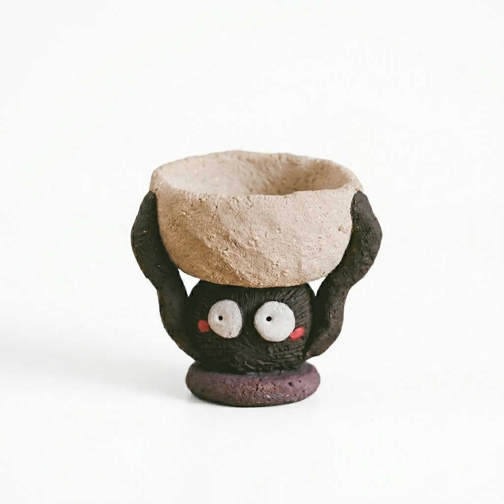 Handmade ceramic soot spirit candle holder featuring a small black soot-spirit character with wide white eyes and rosy cheeks, holding a rustic beige bowl overhead, shown against a white background.