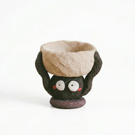 Handmade ceramic soot spirit candle holder featuring a small black soot-spirit character with wide white eyes and rosy cheeks, holding a rustic beige bowl overhead, shown against a white background.
