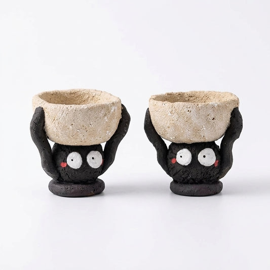 Handmade ceramic soot spirit candle holder shown in two views, featuring black soot spirit figures with wide eyes holding rustic clay bowls on their heads against a clean white background.