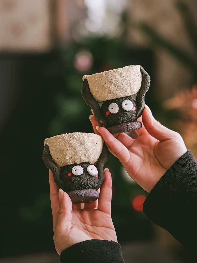 Two handmade ceramic Soot Spirit candle holders held in hands, showing their textured black bodies and cream tops.
