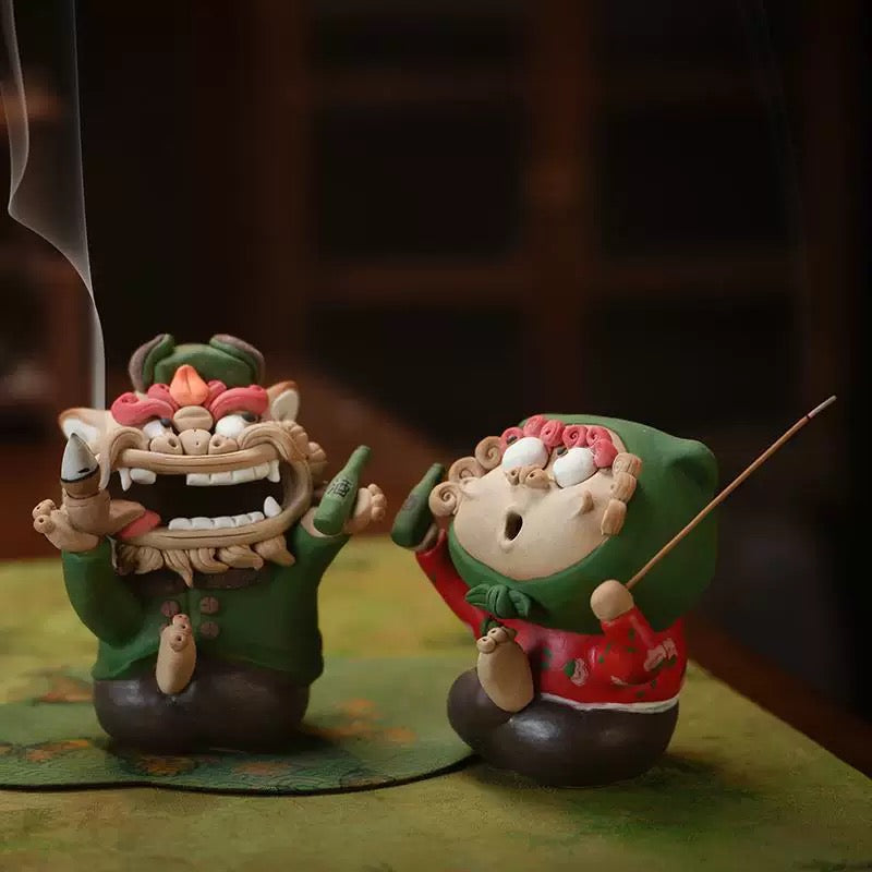 Handmade quirky ceramic incense holder set featuring a green lion backflow burner with smoke and a red figure stick holder on a tabletop in a warm indoor setting