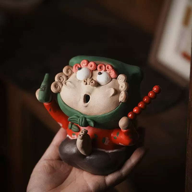 Handmade quirky ceramic incense holder shaped like a red figure holding a bottle and skewer, shown held in hand in a warm indoor setting