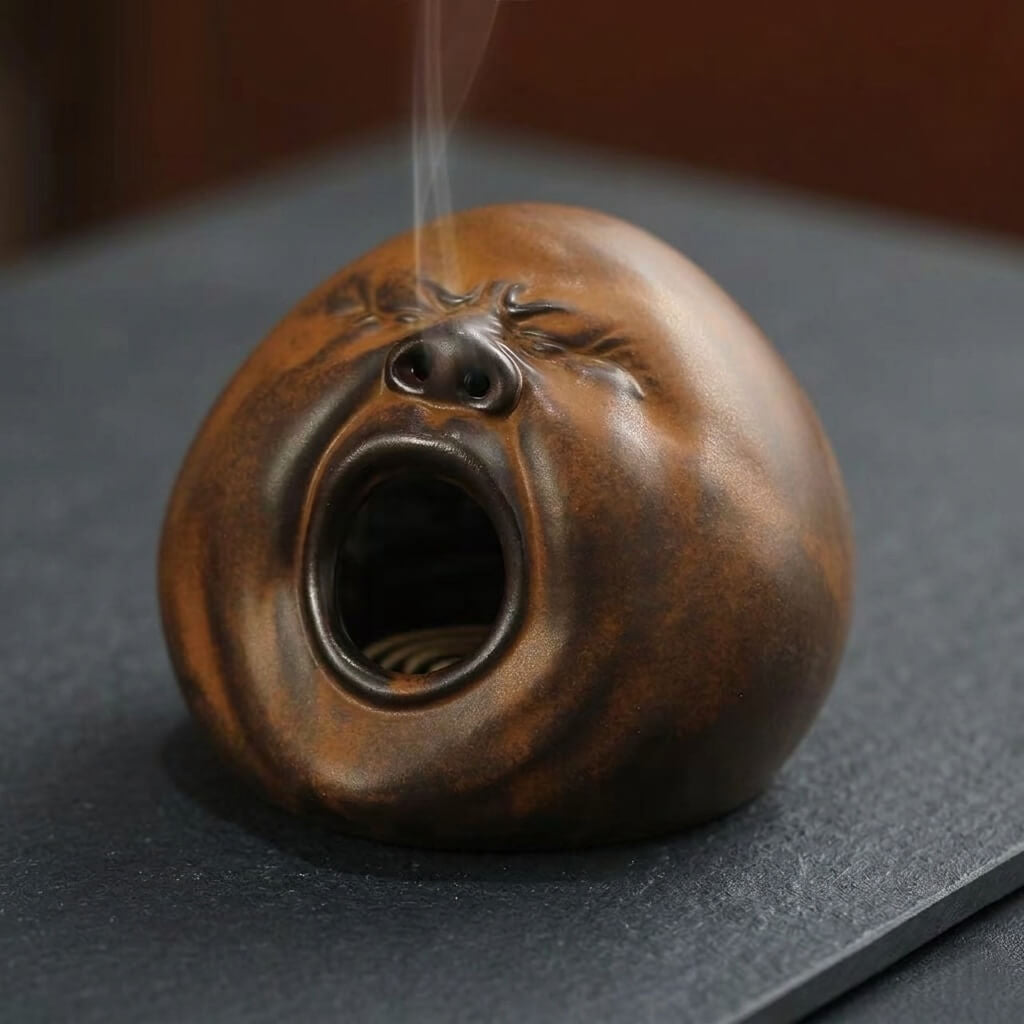 Handmade yawning face incense burner with rounded ceramic design and smoke rising from the top