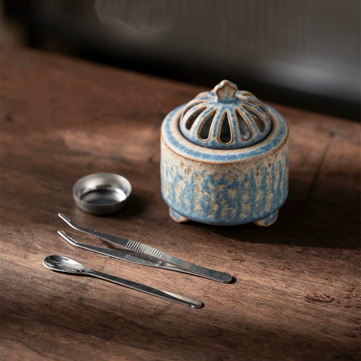 Handmade blue ceramic electric incense burner with stainless steel incense cup, tweezers and spoon laid out on a wooden table.