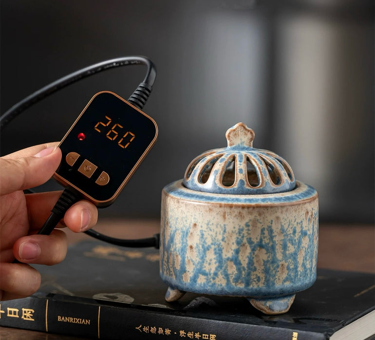 Hand holding a digital temperature controller set to 260 beside a handmade blue ceramic electric incense burner placed on stacked books.